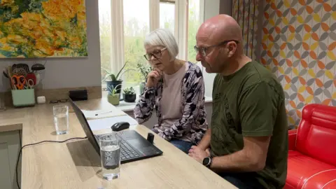 Two people sit at a kitchen counter looking at a laptop, with papers and glasses of water nearby. The room has bright patterned décor, large windows and several plants on the windowsill.
