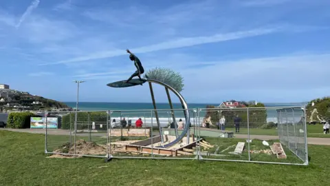 A bronze statue of a surfer behind metal fencing, in its position overlooking a beach and some people walking behind