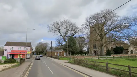 Google Google map view of the road through West Winch. There is a road and on the left side a pink house and the right hand side a church and grassed field. 