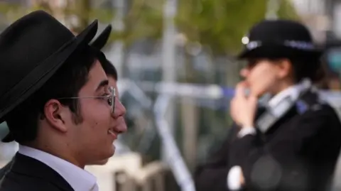 A side view of two people looking over a police cordon in Golders Green. A police officer can be seen blurred in the background.