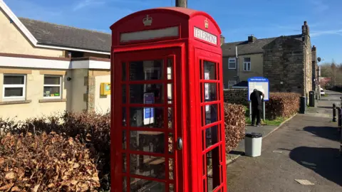 An image of the red phone box on Church Street in Hapton, in the background is the church to the left and to the right a man in black is gardening.