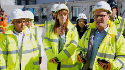 PA Media Mayor of London Sadiq Khan stands next to housing secretary Angela Rayner and prime minister Keir Starmer at a building site - all three are wearing high-viz jackets, hard hats, goggles and work gloves. 