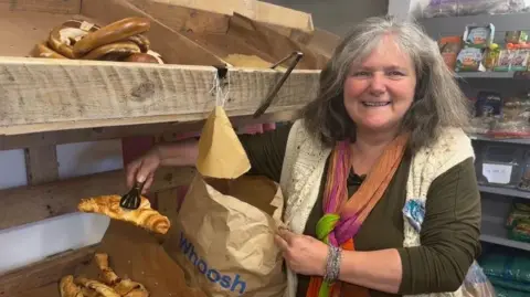 A woman with greying shoulder length hair is standing next to wooden shelves that are holding pastries. She is lifting a croissant into a brown paper bag. She is wearing a brown and white top with a colourful scarf. She is smiling at the camera.