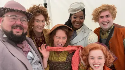 Demitri Lampra A gathering of people in brightly coloured Victorian-style costumes with wavy hair and hats