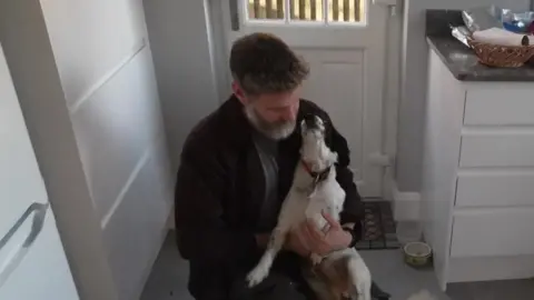 A man wearing an open shirt and a t-shirt is crouched down on a kitchen floor with a black and white dog in his arms. He is looking at the dog.
