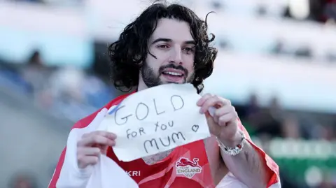England's Zachary Shaw with a St George flag wrapped around him shows a sign saying 'Gold for you Mum' 