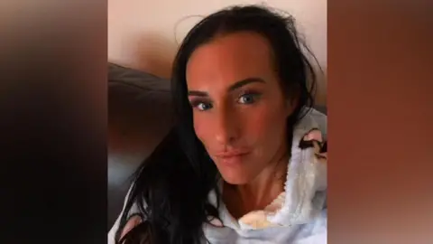 A selfie of Lisa Denton - she has long dark hair and stares into the camera. She is pictured sitting on a sofa, wearing a fluffy hoodie decorated with dogs. 