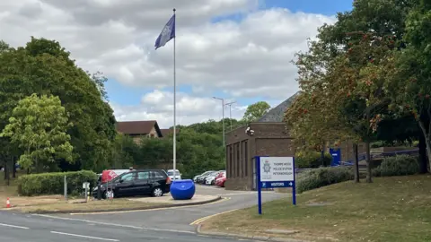 The entrance to Thorpe Wood police station, with a white board with a blue "Thorpe Wood" sign and a blue flag. There are several cars parked to the side and front of the building