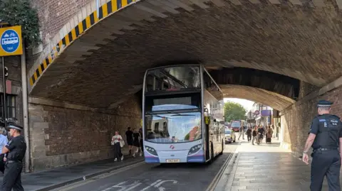 Tracey Franklin Un autobús de dos pisos de color malva que pasa por debajo de un túnel bajo, un policía está en el camino a la derecha y otro a la izquierda de la imagen.