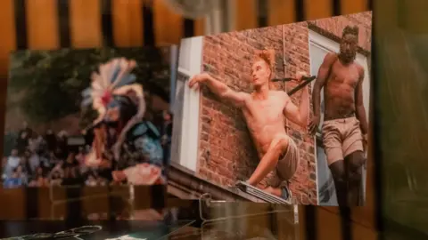 Simon Shaw Two images are displayed side by side in the exhibition. The one on the left shows a man crouching on a small platform between two windows of a red-brick building with his arm outstretched. Another man is standing by the window behind him. The second image is out of focus.