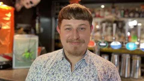 James Stone, a bar manager in Cirencester. He is wearing a flowery blue and white shirt. He has brown hair and has a small beard. Behind him is a well stocked bar full of bottles and glasses.