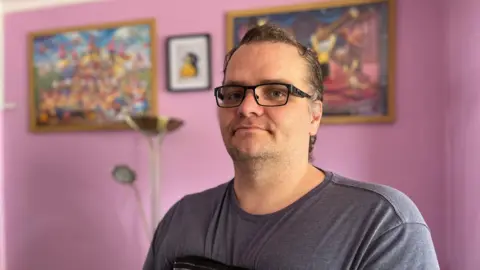 Neil Ferriby. He has short dark hair and wears black rimmed glasses. He is wearing a grey t-shirt and has a black elasticated support bandage around his chest. He is standing in front of a pink wall with three framed prints on it.