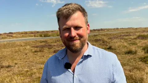 A head and shoulders shot of Ian. He is wearing a blue shirt and has short mousy brown hair. He is stood in front of a field separated by a small road.
