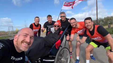 A group of men smile in a selfie wearing orange t-shirts surrounding Jon in a wheelchair.