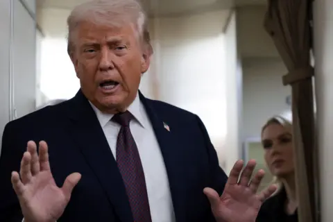 Reuters President Trump stood in the aisle of Air Force One wearing a dark suit and dark red and blue chequered tie. He is holding his hands up. He has wispy, light hair. His press secretary Karoline Levitt is stood in the background with a beige curtain to her left in front of her. She has blonde hair.