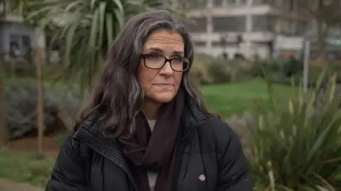 Jacqueline Wilson looks off camera in an outside shot with a hospital building and gardens visible behind her. She is wearing a black coat, dark-rimmed glasses and a dark coloured scarf. She has long dark hair.