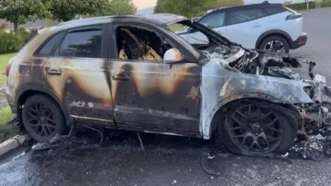 A car is badly burnt with a number of scorch marks.
Several windows have been smashed and the front of the car has melted away.
