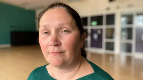 BBC Det Ch Supt Linday Fisher - a woman with tied back dark hair wearing a green top stands in a room with a green wall to her left and a row of windows behind her. The background is blurred.