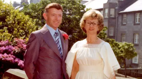 PA Media George Murdoch wearing a grey suit, with a light blue shirt and grey and blue striped tie with wife Jessie, wearing a white dress and glasses. The couple are standing in front of plants, one of which has bright pink flowers, and grey tenement blocks in Aberdeen in 1977.