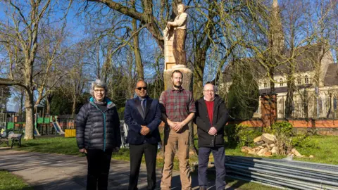 South Swindon Parish Council Four councillors (a woman on the left and three men on her right) are standing in front of a wooden statue in a park, on a sunny day.