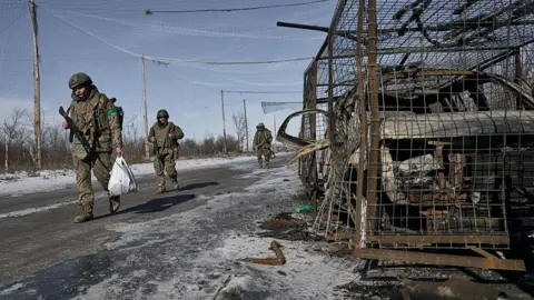 Three soldiers walk on an icy road in their military fatigues carrying weapons and sacks. A burnt out car in a cage is on the side of the road on the right.