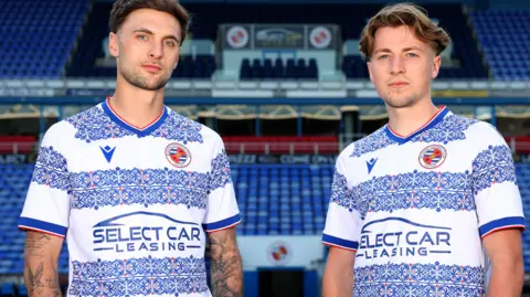 (L-R) Lewis Wing and Charlie Savage smiling at the camera are wearing the new home kit t-shirts. The shirts are blue, white and red with Victorian-inspired flowery patterns.