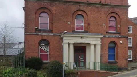 The front of a two-storey brick building with arched windows and a door with white pillars on either side and fencing surrounding it