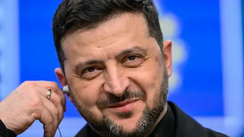 President Zelensky grins as he puts a headphone in his right ear. 