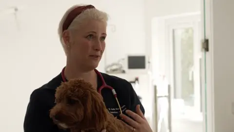 BBC Dr Emily Nightingale is talking her in veterinary surgery. She has short blonde hair in a pixie cut with a red headband on. A stethoscope sits around her neck. A poodle dog sits in front of her on an examination table while she talks.