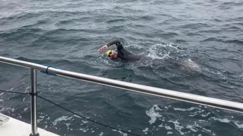 Stewart Douglas One of the swimmers in the sea. He's wearing a yellow swim cap and black goggles and is in a wetsuit. You can see the silver metal railing of the boat.