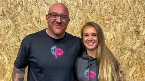 CrossFit Paratus Mr Smart and Ms Blandford standing side by side, posing for the camera in front of a wooden chipping board in the background. They are wearing matching black merch from the gym, with a blue C and a pink P. Mr Smart is bald with a closely shaven beard, wearing dark framed rectangular glasses with a tattoo sleeve on his right arm. His left arm is around Ms Blandford, who has straight blonde hair.