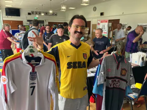 Warren Muggleton/BBC A man wearing a yellow SEGA branded football shirt holds up two sets of shirts on coathangers. He smiles at the camera and has funky yellow glasses and a 70s style moustache. Behind him there are stalls and people milling about.