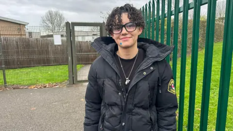 A woman with curly dark hair and a black coat. She is wearing glasses and stood in front of the school gates.