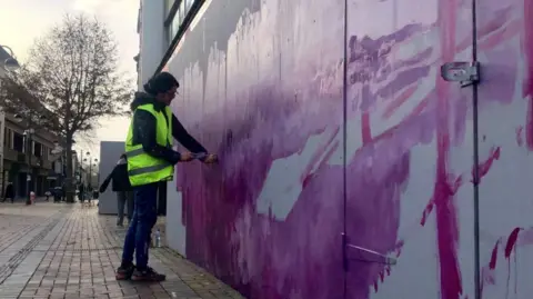Artsite A person standing on a street wearing a high vis jacket painting a wall with purple paint.