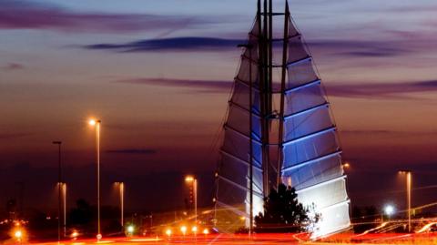 The Tri-Sail sculpture, which has sails like a yacht, pictured during an orange sunset and also showing the lamposts and other lights in the area lit up, creating an orange glow