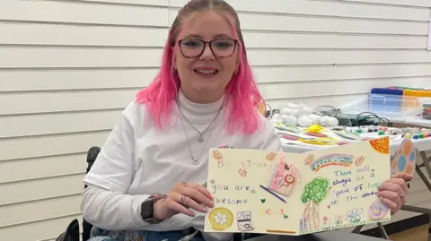 Katie Callaghan is wearing a white roll neck and has pink hair. She is sat at a black table which is covered in handmade cards. She is holding one open towards the camera.