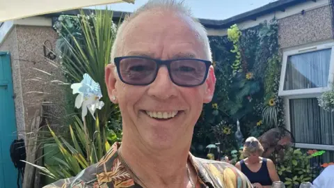 Paul Gregory-Ward Selfie of Paul Gregory-Ward smiling in the sunshine. He is wearing sunglasses and a floral shirt with a bold print and has short white hair