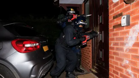 Two police offices holding electric screwdrivers try to get  into a front door of a property