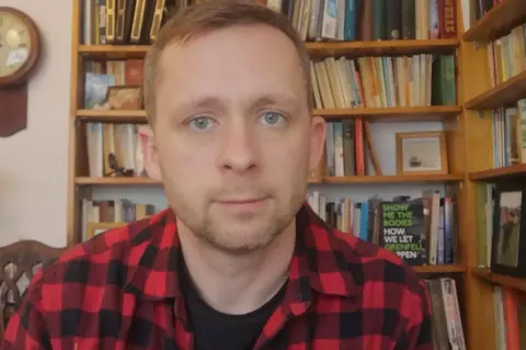 Peter Apps Peter Apps wearing a red and black chequered shirt with a book case filled with books behind him
