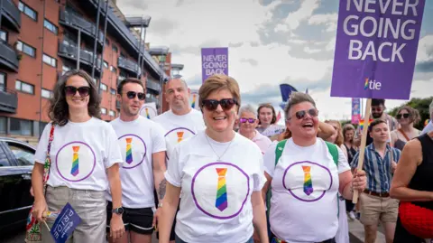 PA Media A group of Pride marchers walk along a city street wearing matching white T-shirts featuring a rainbow-coloured tie logo. One participant carries a large purple sign reading “NEVER GOING BACK”.