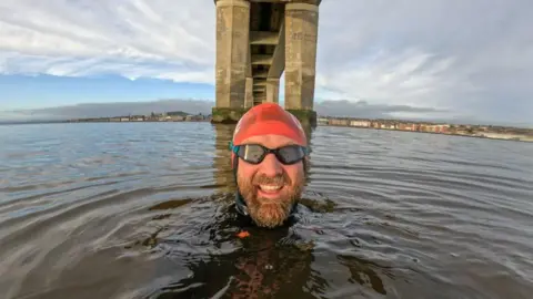 Calum Maclean Image shows swimmer Calum in the water under a stone bridge near Dundee. Only is head is visable above the water. He's wearing a red swim cap and black goggles and has a brown moustache and beard.