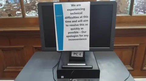  "We are experiencing technical difficulties at this time and will aim to resolve this as quickly as possible - Our apologies for any inconvenience". The screen is on a grey table in front of a wooden background with frosted glass windows.