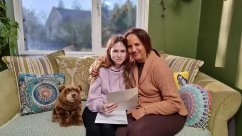 Somerset Council A foster child being hugged by a foster carer on a sofa, smiling to camera and holding a letter