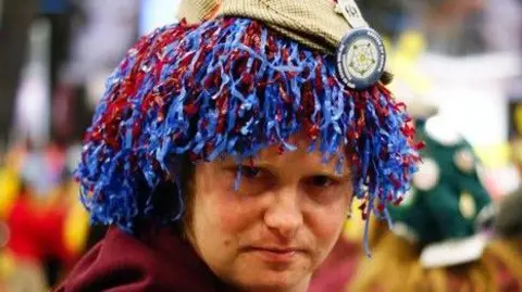 Supplied A man in a red hoodie and blue and red wig looks into the camera. He is wearing a flat cap with a Yorkshire Rose badge and is standing in front of busy crowd. 