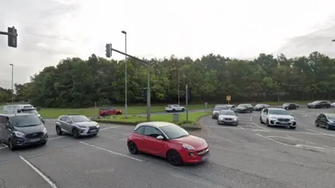 Google Traffic entering onto a roundabout. A row of cars has stopped at traffic lights on the roundabout. There are overhead traffic lights, and trees are visible alongside the road in the background.