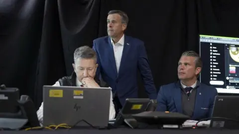 Donald Trump / TruthSocial A photograph posted by U.S. President Donald Trump on his Truth Social account shows U.S. Secretary of Defense Pete Hegseth sitting next to Chairman of the Joint Chiefs of Staff Dan Caine as CIA Director John Ratcliffe stands next to a screen showing posts on the X.com website