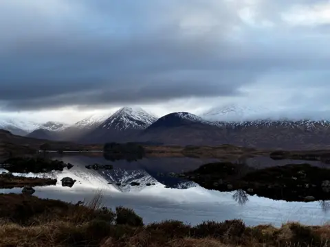 Pete Cocking A calm loch reflects a line of snow‑dusted mountains beneath a moody, overcast sky. 