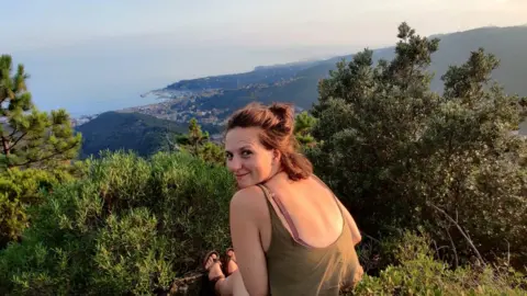 Megan Morgan Megan Morgan sitting on a mountain, looking over her shoulder at the camera. In the distance you can see shrubbery and the sea.