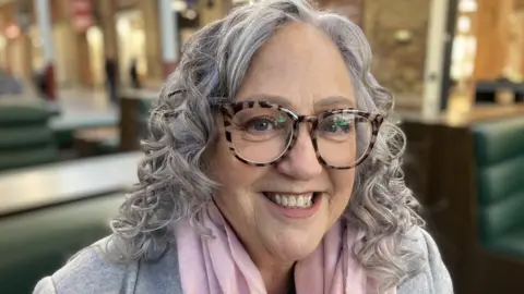 BBC Debbie Brown sat smiling wearing glasses and a pink scarf