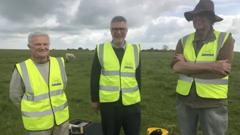 BBC Three archaeologists standing in a field wearing hi-vis jackets